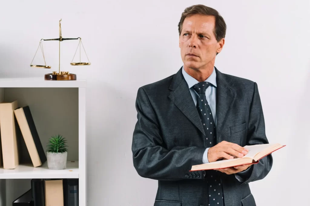 Advogado de terno com um livro, ao lado da balança da justiça e livros jurídicos
